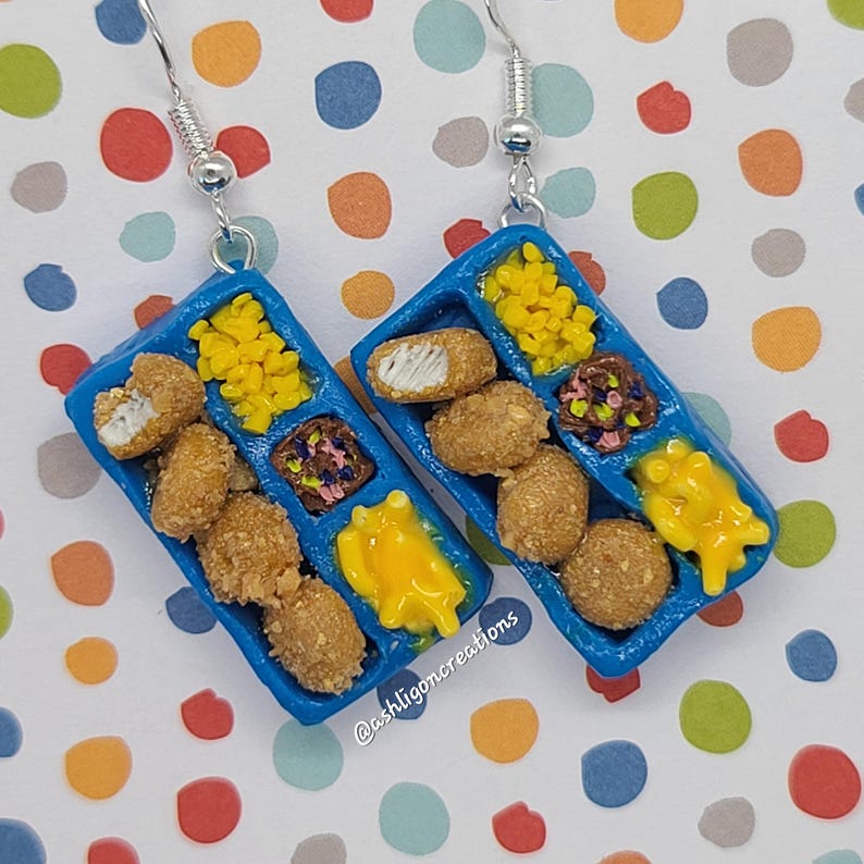 May include: A pair of blue tray earrings with a miniature depiction of a chicken nugget meal. The tray is filled with chicken nuggets, mac and cheese, and a side of fries. The earrings are made of polymer clay and have silver hooks.