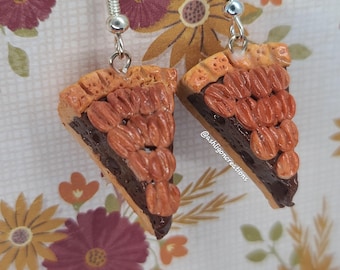 May include: A pair of handmade polymer clay earrings shaped like slices of pecan pie. The earrings are brown, orange, and tan with a chocolate filling and pecan topping. The earrings are on a white background with a floral pattern.