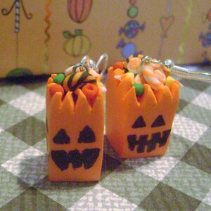 May include: A pair of Halloween-themed earrings featuring orange pumpkin-shaped containers filled with candy corn and other sweets. The earrings are made of polymer clay and have silver hooks.