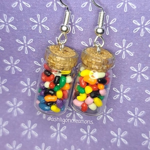 May include: A pair of novelty earrings featuring miniature glass jars filled with colorful jelly beans. Each jar has a cork stopper and is suspended from a silver-tone hook. The background is a light purple with a white floral pattern. The text "@ashligoncreations" is visible.