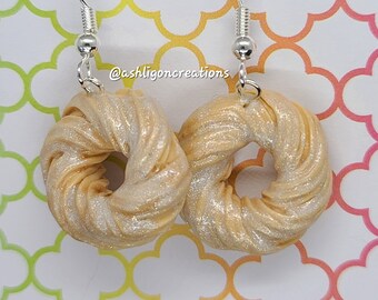 May include: A pair of gold and white donut-shaped earrings with silver hooks. The earrings are made of polymer clay and have a sparkly finish. The earrings are a fun and unique way to add a touch of whimsy to any outfit.