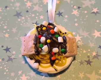 May include: A white ceramic ornament shaped like a plate with a yellow spaghetti-like food topped with chocolate sauce, marshmallows, and colorful candies.