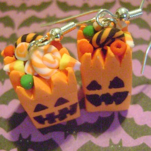 May include: A pair of Halloween-themed earrings featuring orange pumpkin-shaped containers filled with colorful candy, including striped candy canes, orange and yellow candy corn, and black and white striped candy. The earrings are made of polymer clay and have silver hooks.