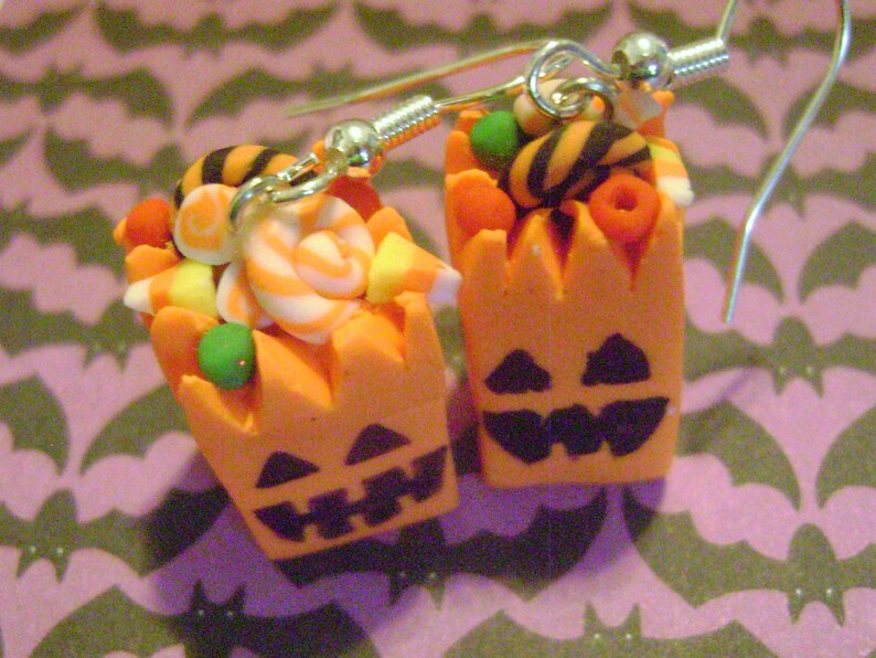May include: A pair of Halloween-themed earrings featuring orange pumpkin-shaped containers filled with colorful candy, including striped candy canes, orange and yellow candy corn, and black and white striped candy. The earrings are made of polymer clay and have silver hooks.