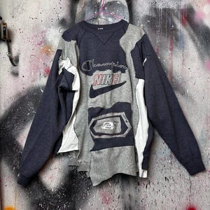 May include: A patchwork sweatshirt in shades of gray and navy blue, featuring the Champion and Nike logos. The design incorporates various fabric panels and visible stitching, creating a unique, deconstructed aesthetic. The sweatshirt is hanging against a textured wall.