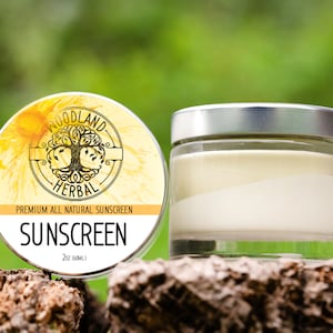 May include: A round jar of Woodland Herbal sunscreen with a yellow and green label. The label says "Premium All Natural Sunscreen" and "Sunscreen" with a tree logo. The jar is sitting on a piece of tree bark.