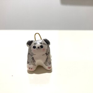 Puede incluir: Una peque&ntilde;a figurita de cer&aacute;mica de un perro con pelaje gris y blanco. El perro est&aacute; sentado con las patas metidas debajo del cuerpo y tiene una expresi&oacute;n linda y caricaturesca.