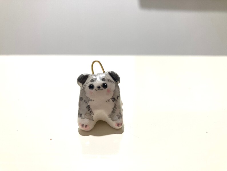 Puede incluir: Una peque&ntilde;a figurita de cer&aacute;mica de un perro con pelaje gris y blanco. El perro est&aacute; sentado con las patas metidas debajo del cuerpo y tiene una expresi&oacute;n linda y caricaturesca.