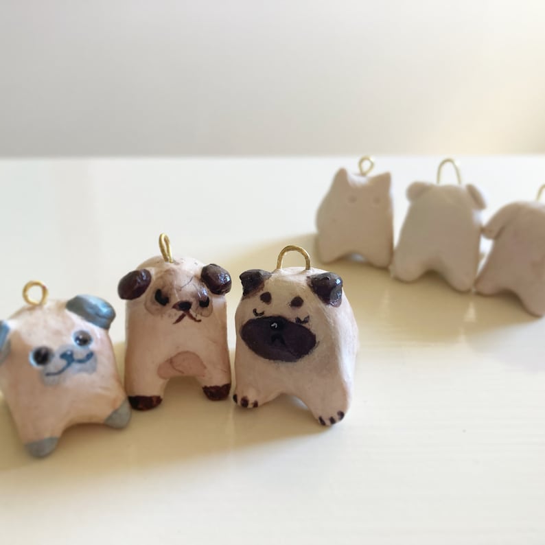 Puede incluir: Cinco peque&ntilde;os adornos de cer&aacute;mica de perro pug con lazos dorados para colgar. Los pugs est&aacute;n pintados en tonos marrones y blancos con ojos y narices negros.