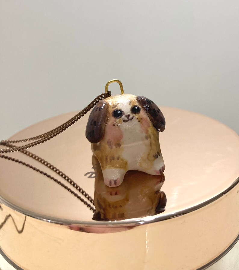 Puede incluir: Un dije de perro de cer&aacute;mica marr&oacute;n y blanco con ojos negros y una cadena de oro. El perro est&aacute; sentado y tiene una expresi&oacute;n feliz.