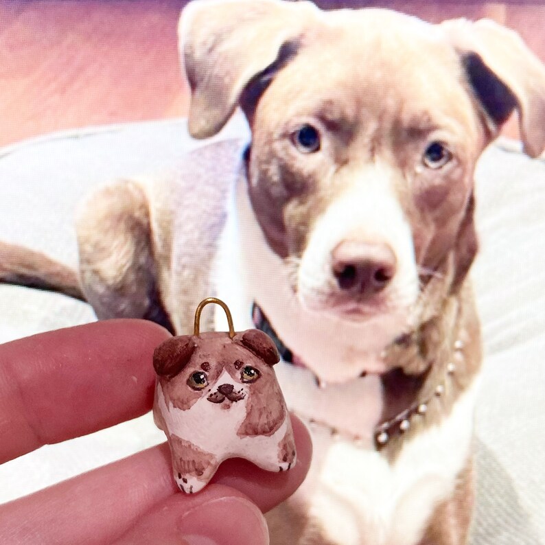Puede incluir: Una peque&ntilde;a figurita de perro de cer&aacute;mica marr&oacute;n y blanca con un lazo dorado para colgar. La figurita tiene una nariz marr&oacute;n, ojos marrones y un pecho blanco.