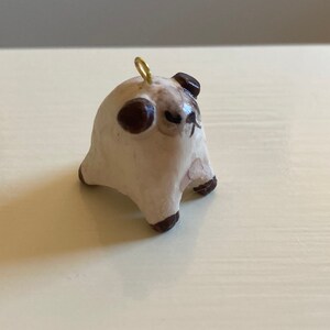 Puede incluir: Una peque&ntilde;a figurita de cer&aacute;mica blanca de un carlino con orejas marrones y un lazo dorado en la parte superior de su cabeza. El carlino est&aacute; sentado con las patas metidas debajo de su cuerpo y tiene una expresi&oacute;n ligeramente gru&ntilde;ona.
