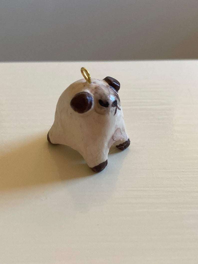 Puede incluir: Una peque&ntilde;a figurita de cer&aacute;mica blanca de un carlino con orejas marrones y un lazo dorado en la parte superior de su cabeza. El carlino est&aacute; sentado con las patas metidas debajo de su cuerpo y tiene una expresi&oacute;n ligeramente gru&ntilde;ona.