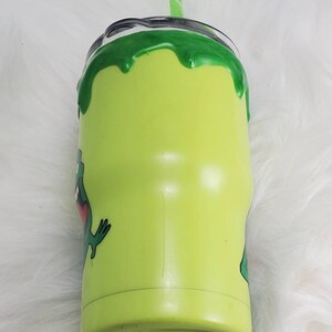 May include: A green stainless steel tumbler with a green slime design and a cartoon frog graphic. The tumbler has a straw and a lid.