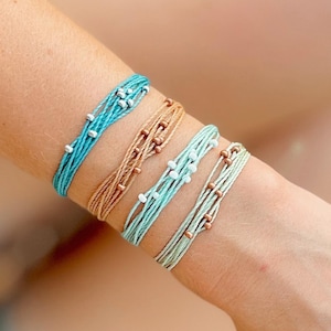 May include: Five colorful string bracelets with small beads are stacked on a wrist. The bracelets are in shades of turquoise, tan, and green. The beads are white and copper-colored. The bracelets are made of multiple strands of string.