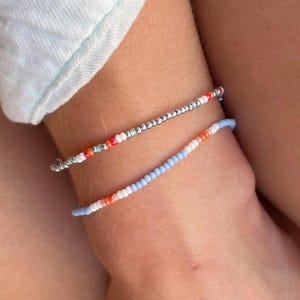 Puede incluir: Dos pulseras de cuentas se llevan en una muñeca. Una pulsera es plateada con cuentas rojas y naranjas. La otra pulsera es azul claro con cuentas blancas y naranjas. Las pulseras están en la muñeca de una persona.