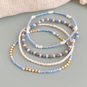 Könnte beinhalten: Vier Armbänder aus Perlen in Blau-, Weiß- und Goldtönen. Die Armbänder bestehen aus kleinen, runden Perlen und sind übereinander gestapelt. Im Hintergrund befindet sich ein helles Tuch mit Spitzenbesatz.