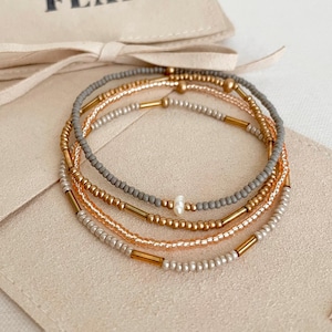 May include: A set of four beaded bracelets in gold, gray, and silver tones. The bracelets are displayed on a beige pouch with the text "CAITLYN CLARE FLAIR". The beads are small and delicate, with a mix of round and tube-shaped beads.