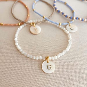 Può includere: Cinque braccialetti di perline in vari colori: pesca, marrone, azzurro e bianco. Ogni braccialetto presenta un ciondolo rotondo bianco con un'iniziale dorata. I braccialetti sono infilati con piccole perline e dettagli dorati, creando un accessorio delicato e personalizzato.