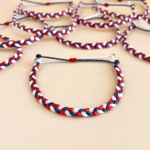 May include: Braided bracelets in red, white, and blue. The bracelets are made of woven cord, with a repeating pattern of the three colors. The bracelets are displayed on a light beige surface. The bracelets are a patriotic theme.