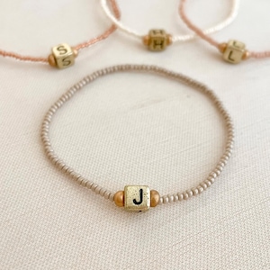 Puede incluir: Pulseras de cuentas en tonos rosa, crema y topo. Cada pulsera presenta un pequeño cubo dorado con una letra. Las pulseras se exhiben sobre una superficie neutra. Las letras visibles son S, H, L y J.
