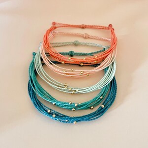 K&ouml;nnte beinhalten: Stapel bunter Freundschaftsarmb&auml;nder. Die Armb&auml;nder bestehen aus geflochtenem Kordel in den Farbt&ouml;nen Koralle, Rosa, T&uuml;rkis und Blau. Jedes Armband hat kleine goldfarbene Perlen. Die Armb&auml;nder sind &uuml;bereinander gestapelt.
