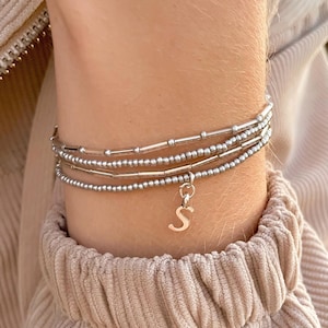 Puede incluir: Un conjunto de cuatro pulseras de cuentas plateadas. Una pulsera tiene un pequeño dije plateado con la forma de la letra "S". Las pulseras están hechas de pequeñas cuentas redondas y cuentas cilíndricas. Las pulseras están apiladas.