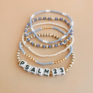 Puede incluir: Conjunto de pulseras de cuentas en tonos azules, blancos y dorados. Una pulsera presenta cuentas cuadradas blancas que deletrean "PSALM 23". Las pulseras están dispuestas sobre una superficie melocotón claro.