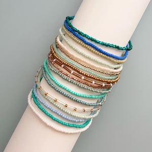 May include: A collection of beaded bracelets in various colors, including green, blue, white, and gold. The bracelets are stacked on a white cylindrical form, showcasing their different designs and textures. The beads appear to be small and delicate.