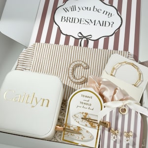 May include: A gift box containing a white jewelry case with the name Caitlyn, a striped pouch with a pearl-embellished letter C, a gold chain bracelet, a satin scrunchie, two gold-flecked hair clips, and bow earrings with pearl accents.