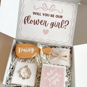 May include: A gift box with the text "Will you be our flower girl?" contains a wooden hairbrush, a bracelet, a hair clip, and a small box. The items are arranged on white paper shreds. The box and items have a pink and white colour scheme.