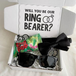 Puede incluir: Una caja de regalo blanca que contiene una pajarita negra, gafas de sol negras, un caramelo Ring Pop verde y dos pequeños recortes negros en forma de anillo. La tapa interior muestra el texto WILL YOU BE OUR RING BEARER? con dos anillos entrelazados.