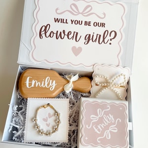 May include: A white gift box with a pink and white floral design, containing a wooden hairbrush, a bracelet, a hair clip, and a small box. The hairbrush is engraved with the name "Emily". The box lid reads "Will you be our flower girl?"