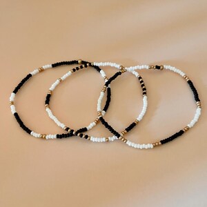 Può includere: Tre braccialetti di perline nere, bianche e dorate. Ogni braccialetto presenta un motivo diverso di perline nere, bianche e dorate. I braccialetti sono esposti su una superficie marrone chiaro. Il testo sull'immagine recita "ONE OF EACH STYLE."