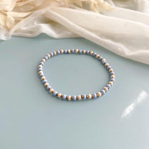Peut inclure: Un bracelet circulaire compos&eacute; de perles alternant un motif r&eacute;p&eacute;titif de petites perles bleu clair, de perles blanches &agrave; la texture marbr&eacute;e et de perles m&eacute;talliques dor&eacute;es, dispos&eacute;es en une s&eacute;quence r&eacute;guli&egrave;re sur toute la longueur du bijou.