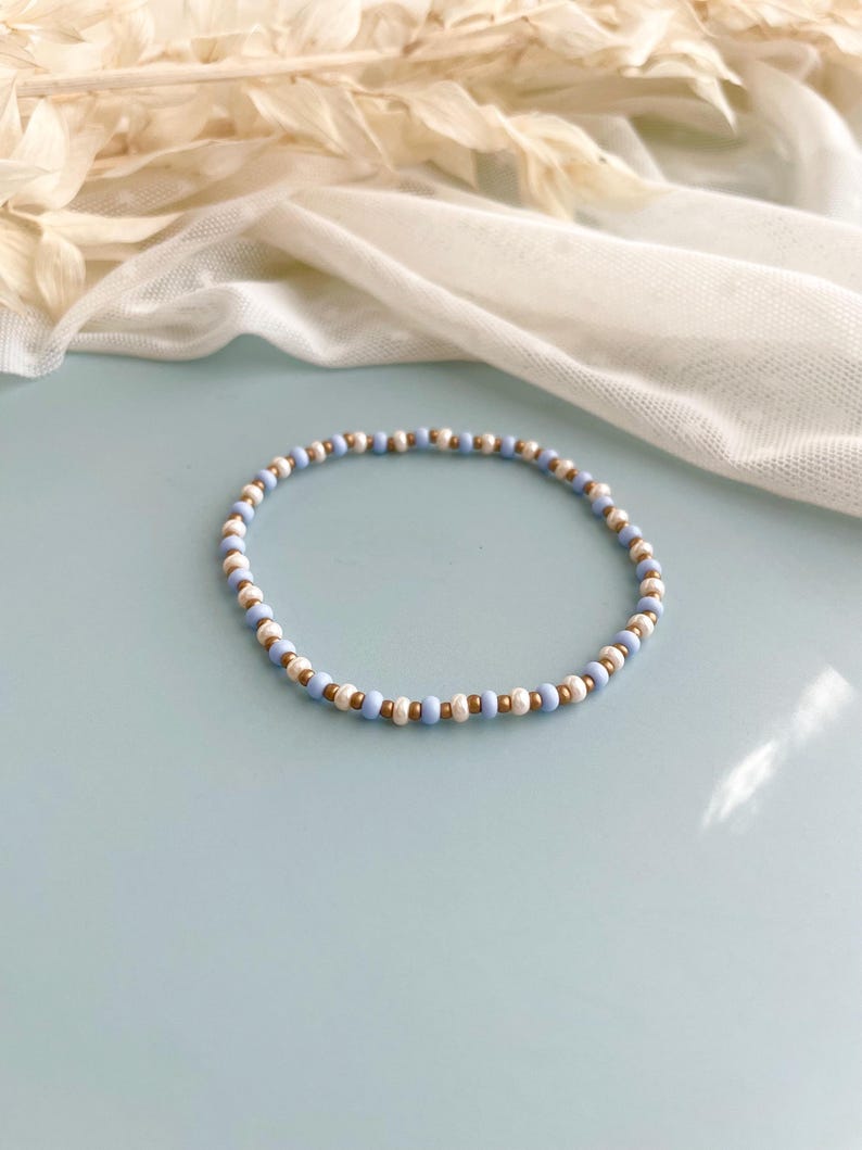 Peut inclure: Un bracelet circulaire compos&eacute; de perles alternant un motif r&eacute;p&eacute;titif de petites perles bleu clair, de perles blanches &agrave; la texture marbr&eacute;e et de perles m&eacute;talliques dor&eacute;es, dispos&eacute;es en une s&eacute;quence r&eacute;guli&egrave;re sur toute la longueur du bijou.