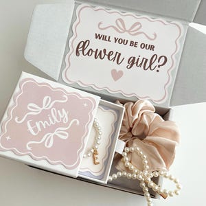 Puede incluir: Una caja de regalo con un diseño rosa y blanco, que contiene una pulsera de perlas con un dije de inicial dorada, una pinza para el pelo de perlas y una scrunchie de satén. La caja incluye una tarjeta que dice "¿Quieres ser nuestra niña de las flores?"