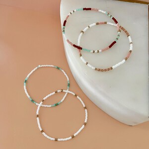 Peut inclure: Plusieurs bracelets de perles de diff&eacute;rentes couleurs. Les bracelets pr&eacute;sentent des perles blanches, roses, turquoise et bordeaux, avec des accents dor&eacute;s. Les bracelets sont dispos&eacute;s sur une surface en marbre blanc sur un fond p&ecirc;che clair.