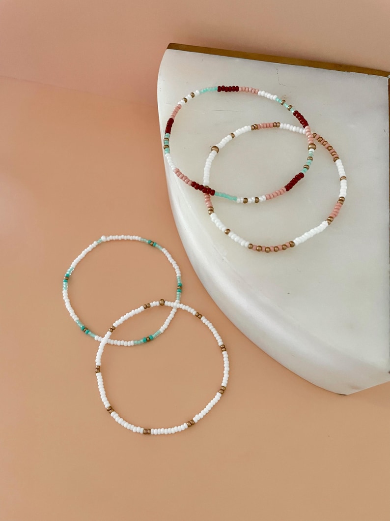 Peut inclure: Plusieurs bracelets de perles de diff&eacute;rentes couleurs. Les bracelets pr&eacute;sentent des perles blanches, roses, turquoise et bordeaux, avec des accents dor&eacute;s. Les bracelets sont dispos&eacute;s sur une surface en marbre blanc sur un fond p&ecirc;che clair.