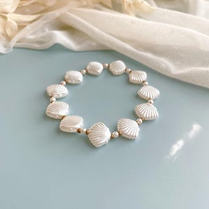 Peut inclure: Un bracelet circulaire compos&eacute; d'une alternance de perles blanches en forme de coquillage et de petites perles intercalaires m&eacute;talliques dor&eacute;es. Les perles coquillages pr&eacute;sentent une texture c&ocirc;tel&eacute;e et une finition brillante.