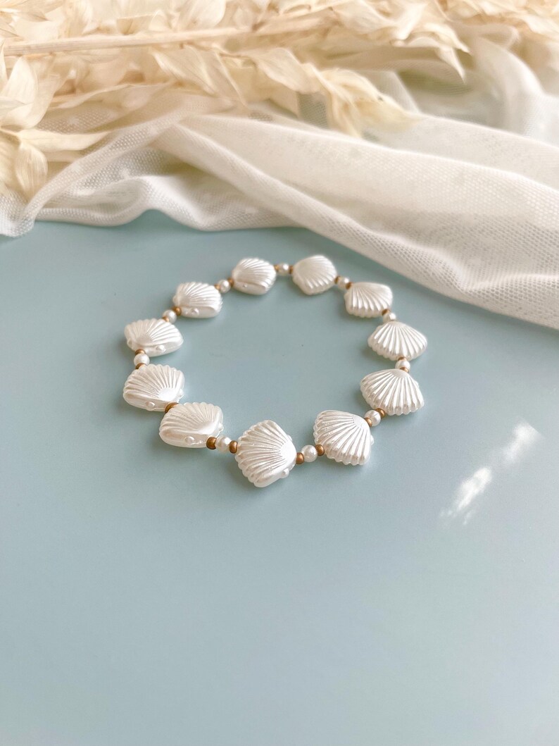 Peut inclure: Un bracelet circulaire compos&eacute; d'une alternance de perles blanches en forme de coquillage et de petites perles intercalaires m&eacute;talliques dor&eacute;es. Les perles coquillages pr&eacute;sentent une texture c&ocirc;tel&eacute;e et une finition brillante.