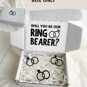 May include: A white cardboard gift box with the text BOX ONLY at the top. The inside lid features the text WILL YOU BE OUR RING BEARER? with a diamond ring icon. The box contains white paper filler and three black diamond ring cutouts.