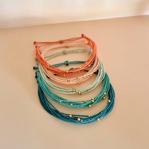 K&ouml;nnte beinhalten: Eine Sammlung farbenfroher Armb&auml;nder aus Schn&uuml;ren in Korallen-, T&uuml;rkis- und Blaut&ouml;nen. Jedes Armband hat mehrere Str&auml;nge, die mit kleinen goldfarbenen Perlen verziert sind. Die Armb&auml;nder sind gestapelt und zeigen ihre verschiedenen Farben und Designs.