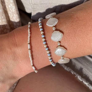 May include: A set of three beaded bracelets. One features small white and gold cylindrical beads. The second consists of alternating light blue, white, and gold spherical beads. The third features white shell-shaped beads with small gold spacers.
