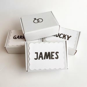 May include: Three white rectangular cardboard gift boxes stacked together. The top box features a black icon of two interlocking rings. The two lower boxes have white scalloped labels with the black printed names GARK, LUCKY, and JAMES.