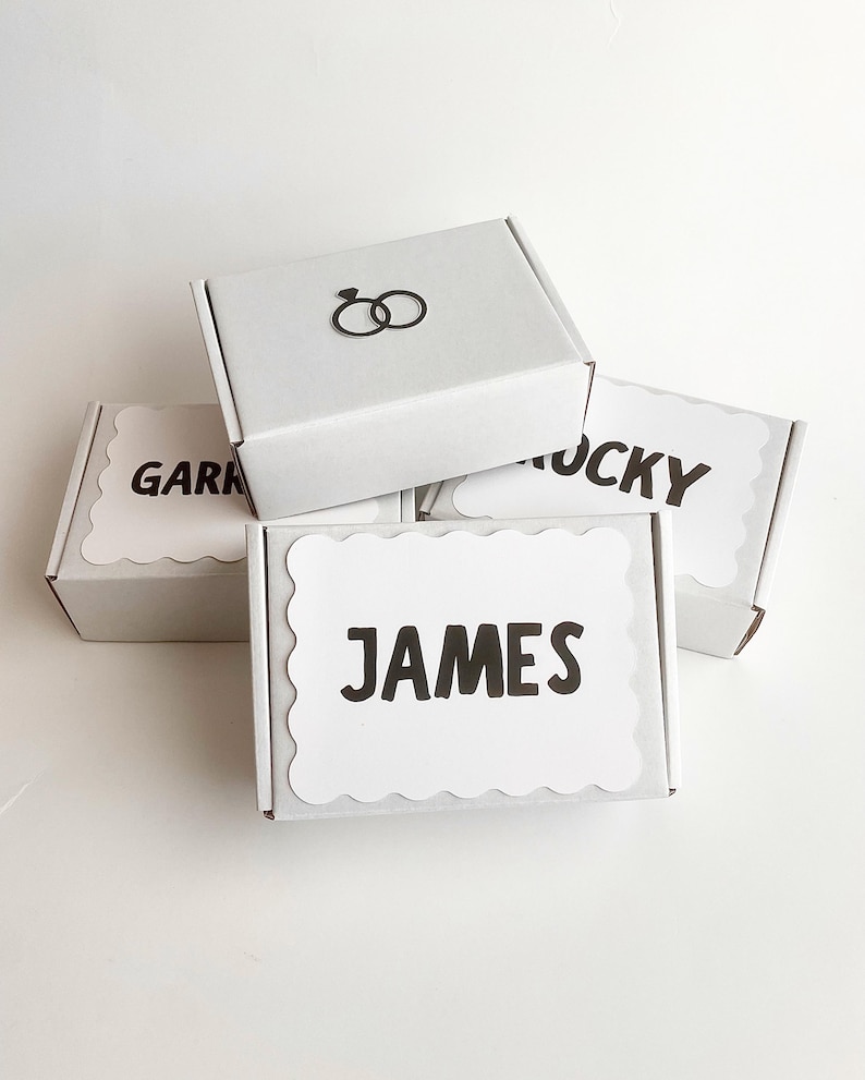 May include: Three white rectangular cardboard gift boxes stacked together. The top box features a black icon of two interlocking rings. The two lower boxes have white scalloped labels with the black printed names GARK, LUCKY, and JAMES.