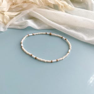 Peut inclure: Un bracelet de perles d&eacute;licat compos&eacute; d'un motif r&eacute;p&eacute;titif de petites perles de rocaille rondes blanc cass&eacute;, altern&eacute;es avec des perles blanches cylindriques plus longues et de minuscules perles d'espacement sph&eacute;riques dor&eacute;es m&eacute;tallis&eacute;es.
