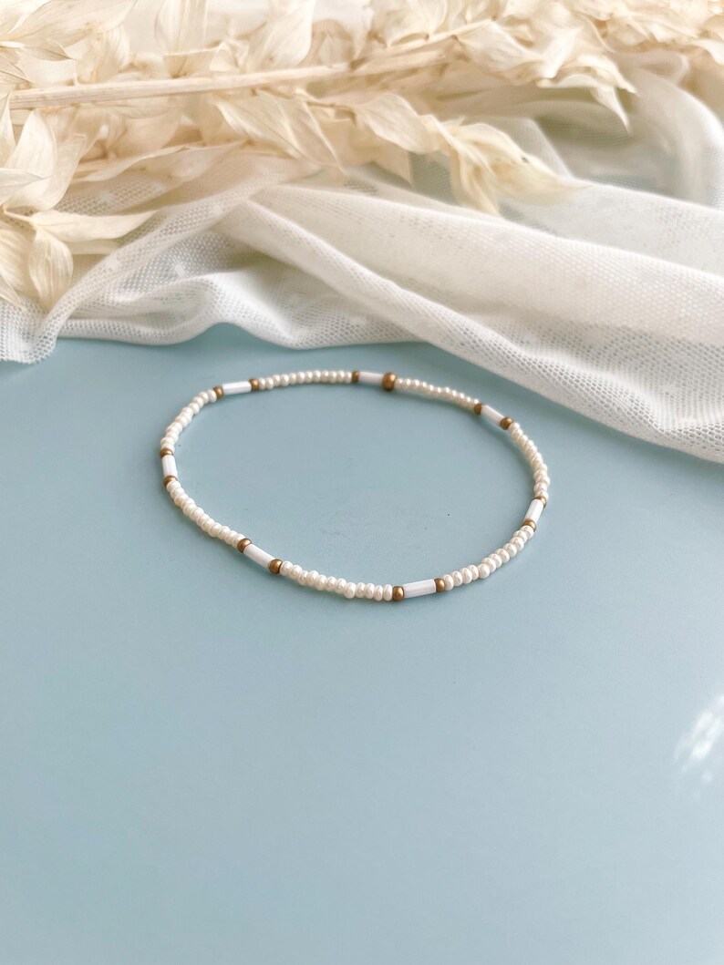 Peut inclure: Un bracelet de perles d&eacute;licat compos&eacute; d'un motif r&eacute;p&eacute;titif de petites perles de rocaille rondes blanc cass&eacute;, altern&eacute;es avec des perles blanches cylindriques plus longues et de minuscules perles d'espacement sph&eacute;riques dor&eacute;es m&eacute;tallis&eacute;es.