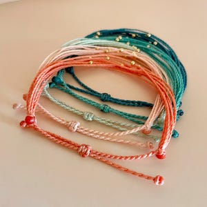 K&ouml;nnte beinhalten: Eine Sammlung farbenfroher Armb&auml;nder aus Schn&uuml;ren in Korallen-, Rosa-, T&uuml;rkis- und T&uuml;rkist&ouml;nen. Jedes Armband hat ein geknotetes Design und kleine goldene Perlen. Die Armb&auml;nder sind in einer geschichteten Anordnung angeordnet.
