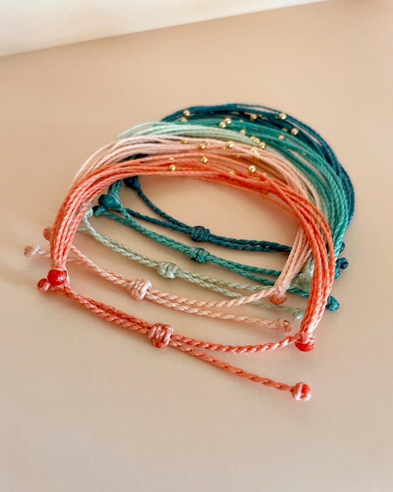 K&ouml;nnte beinhalten: Eine Sammlung farbenfroher Armb&auml;nder aus Schn&uuml;ren in Korallen-, Rosa-, T&uuml;rkis- und T&uuml;rkist&ouml;nen. Jedes Armband hat ein geknotetes Design und kleine goldene Perlen. Die Armb&auml;nder sind in einer geschichteten Anordnung angeordnet.